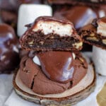 Chocolate marshmallow cookie recipe topped with fudge frosting sitting on wood coaster. More cookies cut in half can be seen in the background.