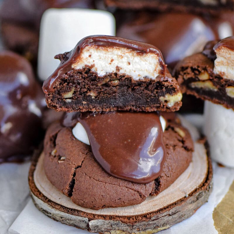 Chocolate marshmallow cookie recipe topped with fudge frosting sitting on wood coaster. More cookies cut in half can be seen in the background.