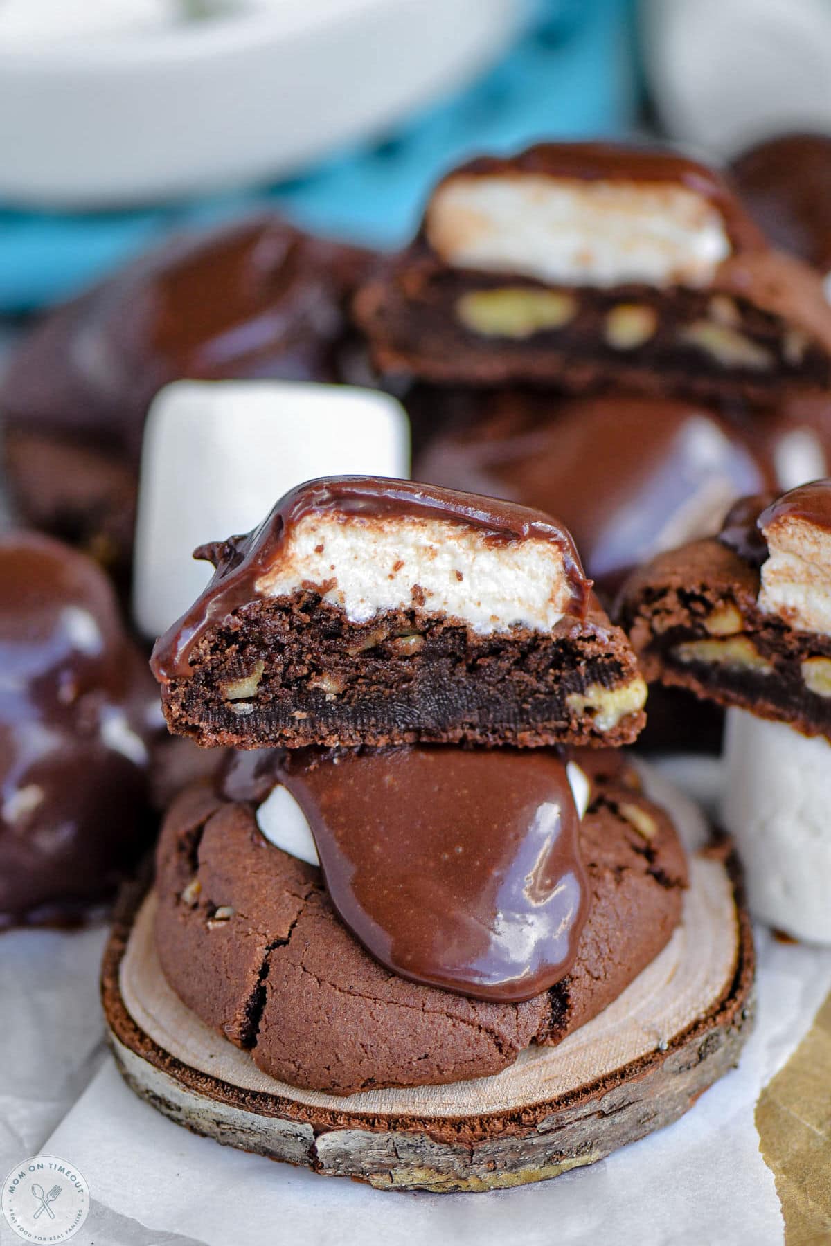 Chocolate cookies topped with marshmallow and a fudge frosting sitting on wood coaster. More cookies cut in half can be seen in the background.