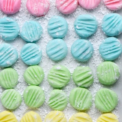 Top down view of recipe for cream cheese mints on parchment paper flattened with the tines of a fork. Four pastel colors are shown: pink, blue, green and yellow in a symmetrical grid layout.