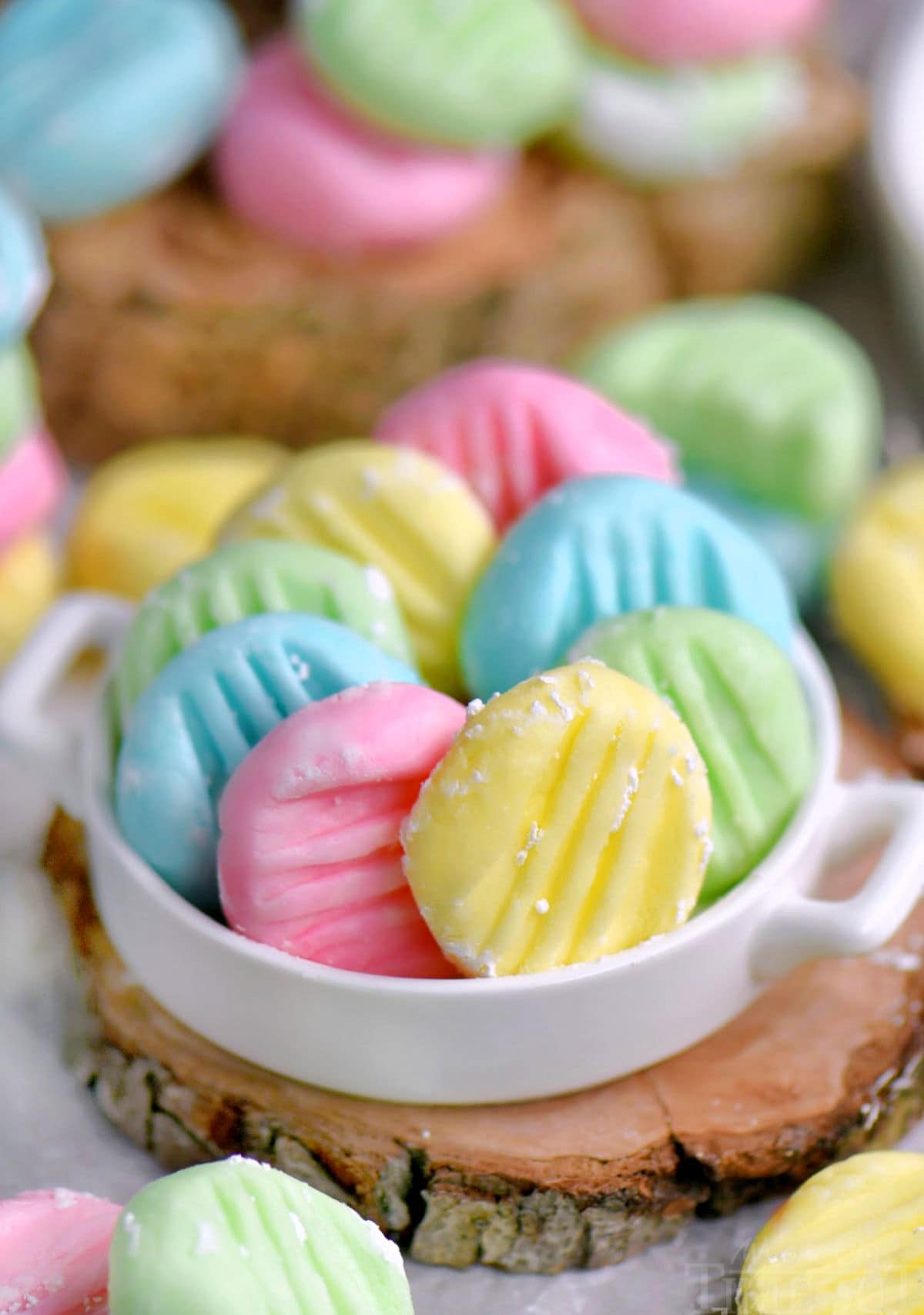 Colorful pastel cream cheese mints in white shallow dish sitting on wood surface.