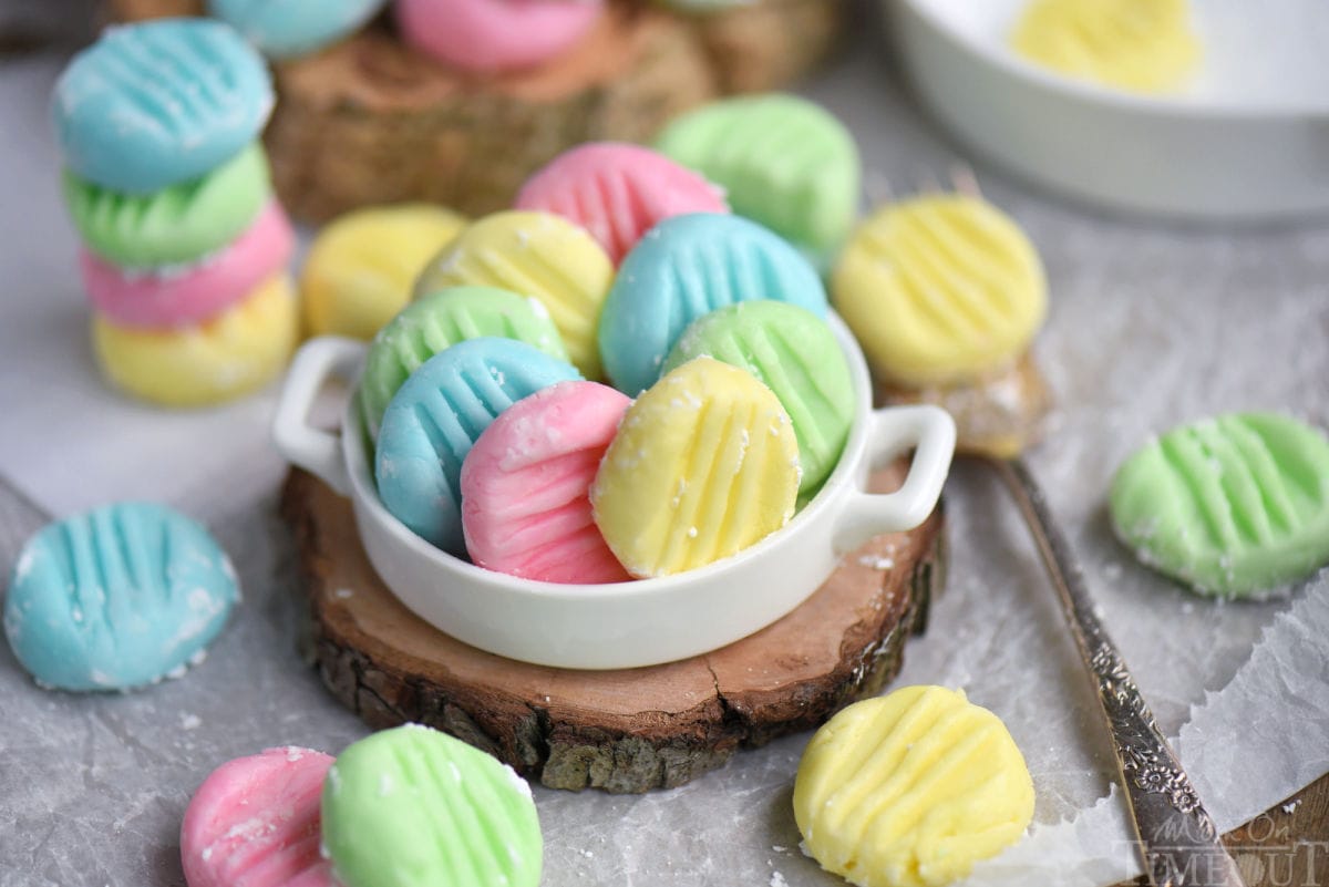 Colorful pastel cream cheese mints in white shallow dish sitting on wood surface. More mints are scattered around the bowl.