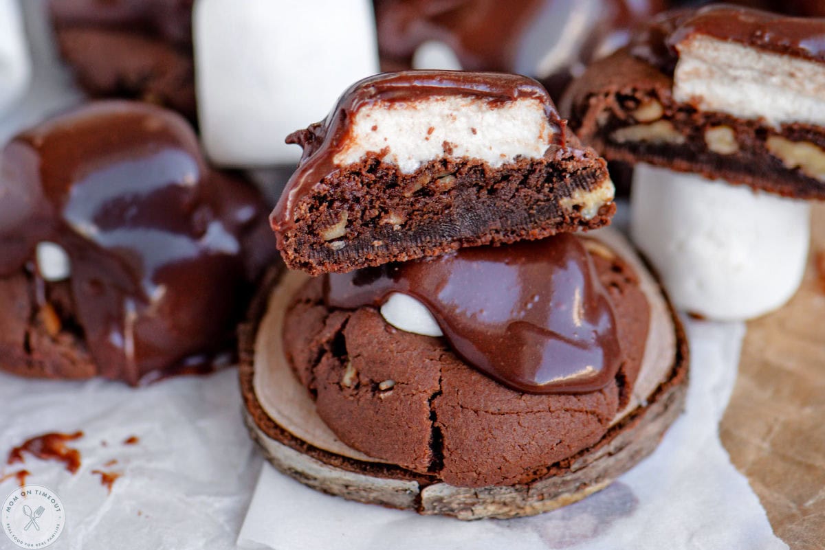Chocolate marshmallow cookies topped with fudge frosting sitting on wood coaster. More cookies cut in half can be seen in the background.