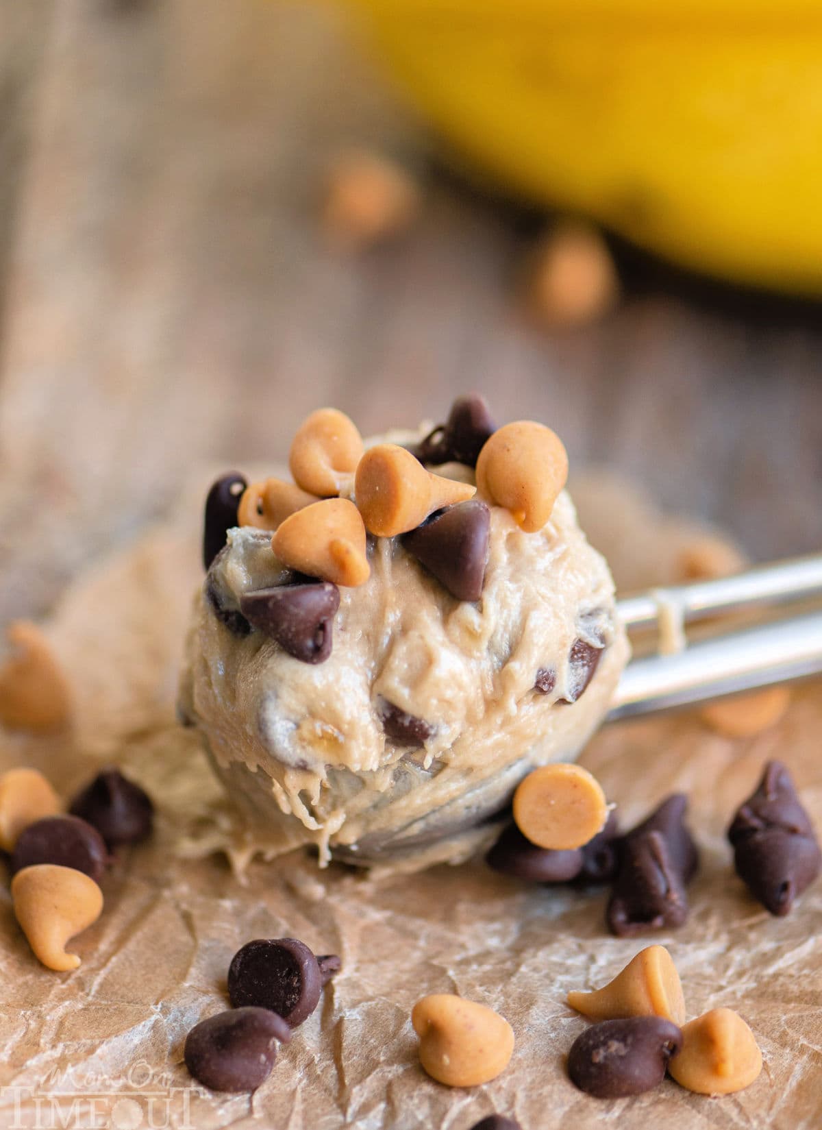 Cookie dough scoop with peanut butter banana chocolate chip cookie dough in it resting on brown parchment with peanut butter and chocolate chips scattered about.