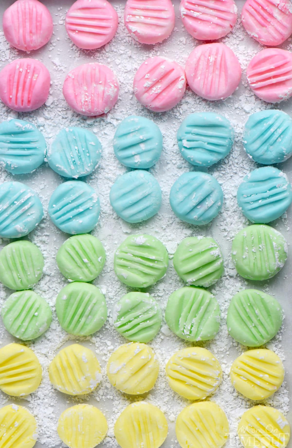 Top down view of recipe for cream cheese mints on parchment paper flattened with the tines of a fork. Four pastel colors are shown: pink, blue, green and yellow in a symmetrical grid layout.