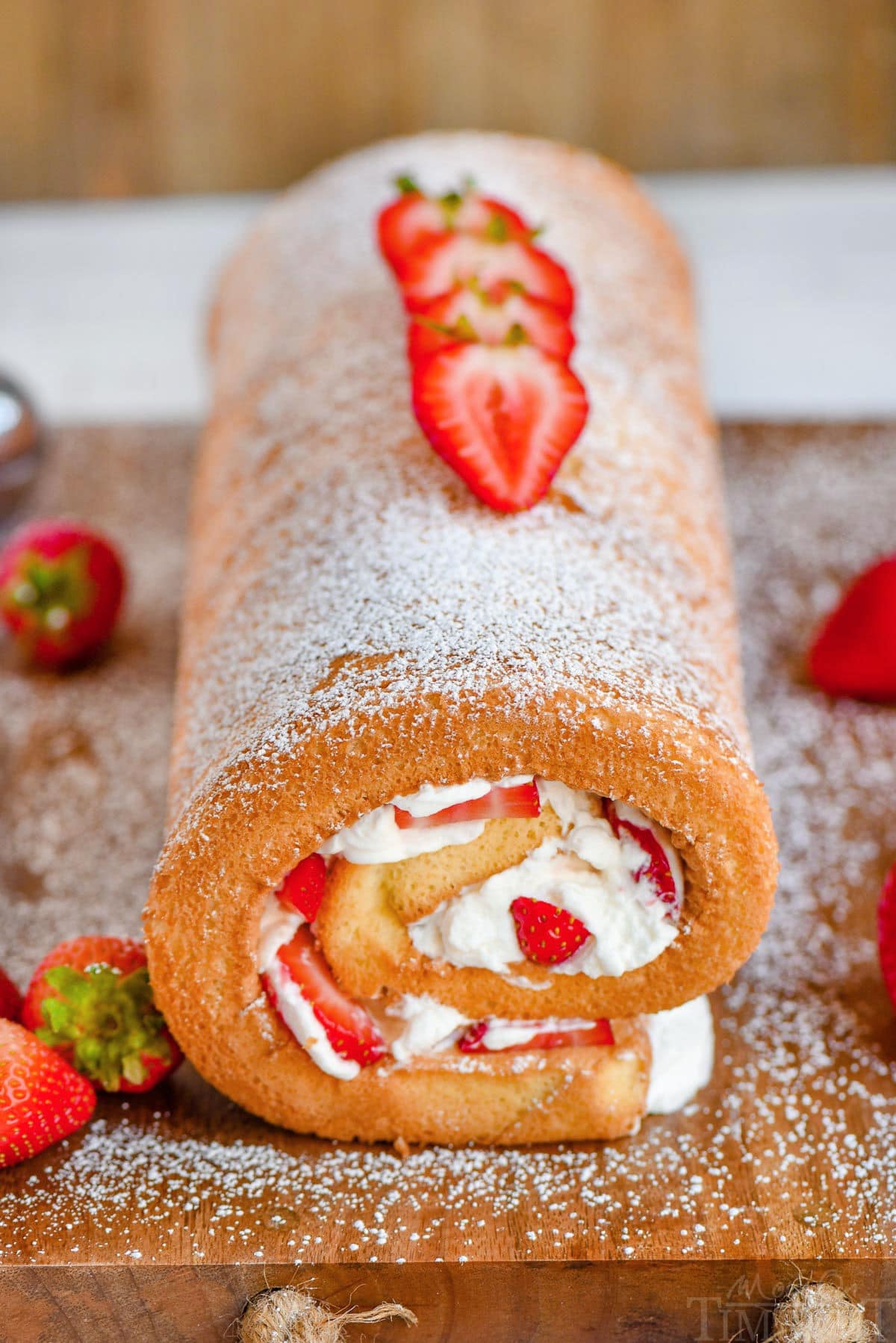 View of whole strawberry shortcake cake roll on wood cutting board ready to be cut and served.