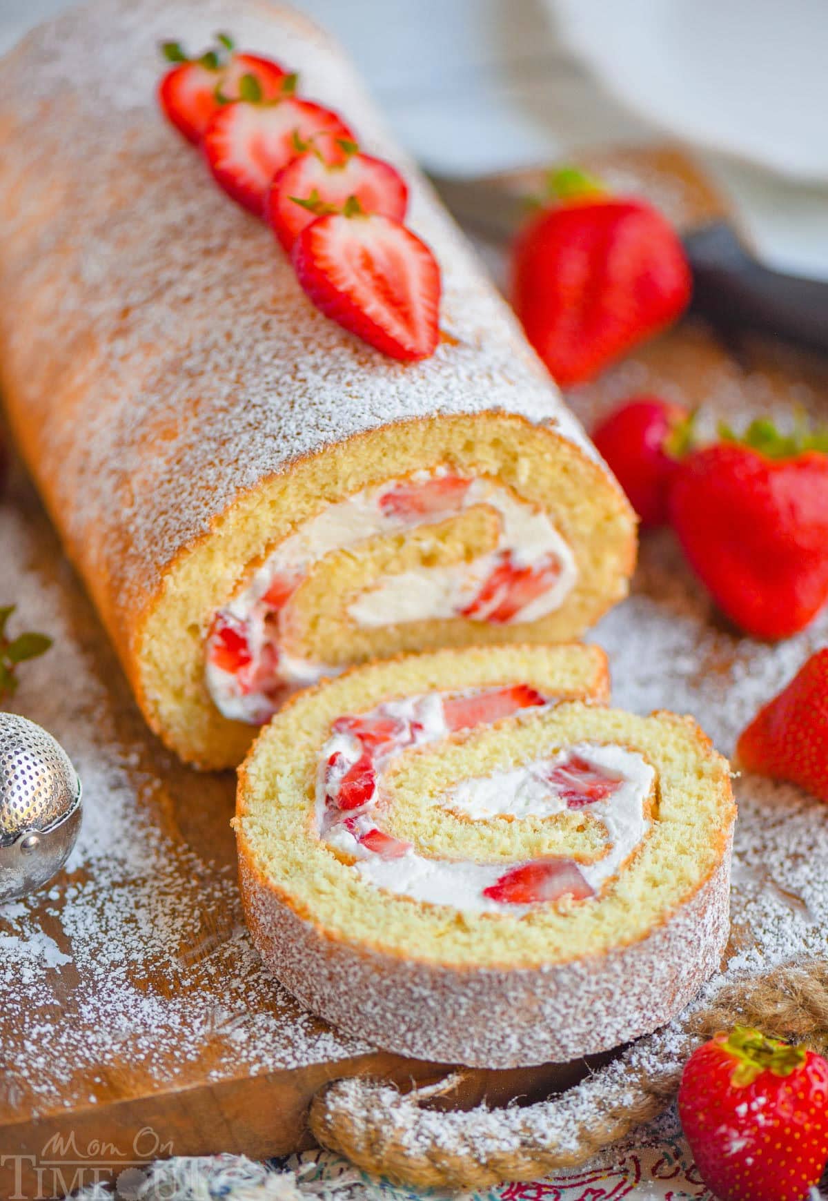 Strawberry Cake Roll with one slice cut off at the end sitting on wood board topped with powdered sugar and fresh strawberries.