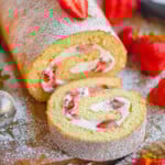 Strawberry Cake Roll with one slice cut off at the end sitting on wood board topped with powdered sugar and fresh strawberries.