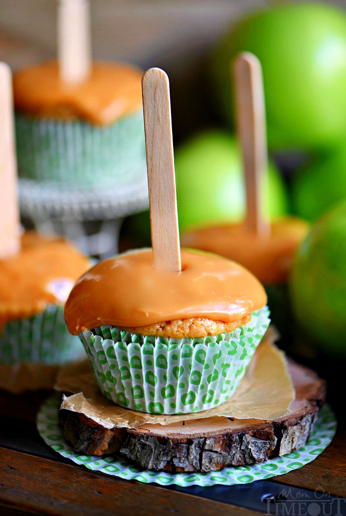 cupcake alle mele guarnito con glassa al caramello e un bastoncino di ghiacciolo inserito nella parte superiore.  Sullo sfondo si possono vedere un altro paio di cupcakes.