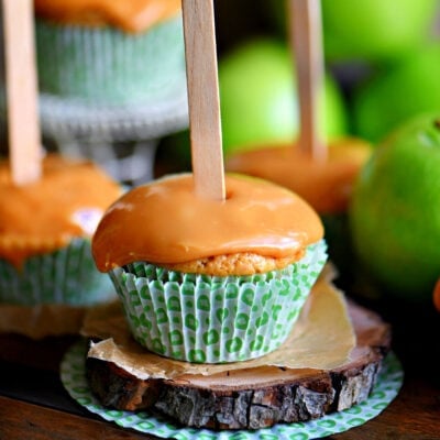 apple cupcake topped with caramel frosting and a popsicle stick inserted into the top. A couple more cupcakes can be seen in the background.