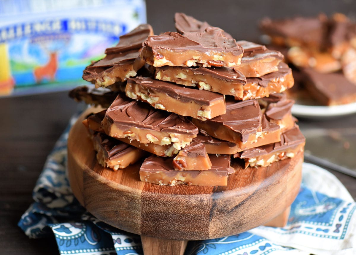 toffee piled high on dark wood round board sitting on blue and white background. box of butter in background.