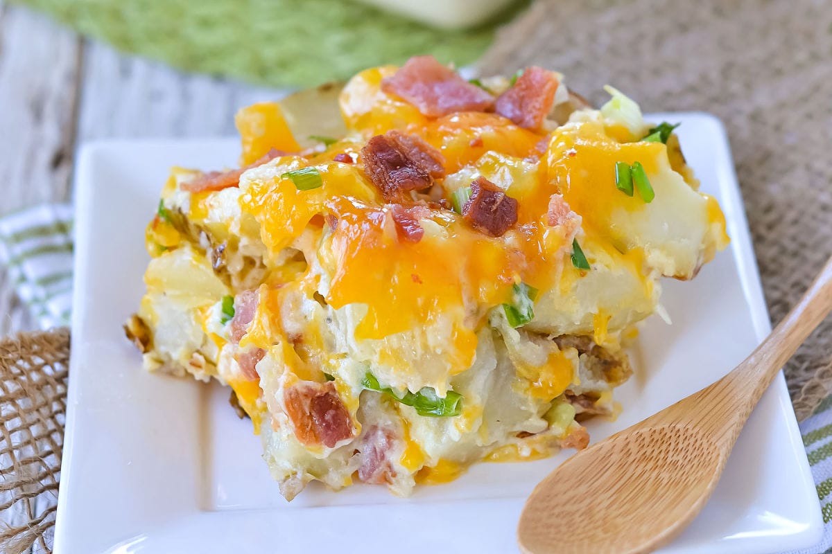 potato casserole topped with shredded sharp cheddar cheese, bacon and green onions sitting on a small white square plate.