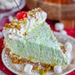 piece of pistachio pie on small white dessert plated garnished with freshly whipped cream, chopped pistachios and a maraschino cherry.