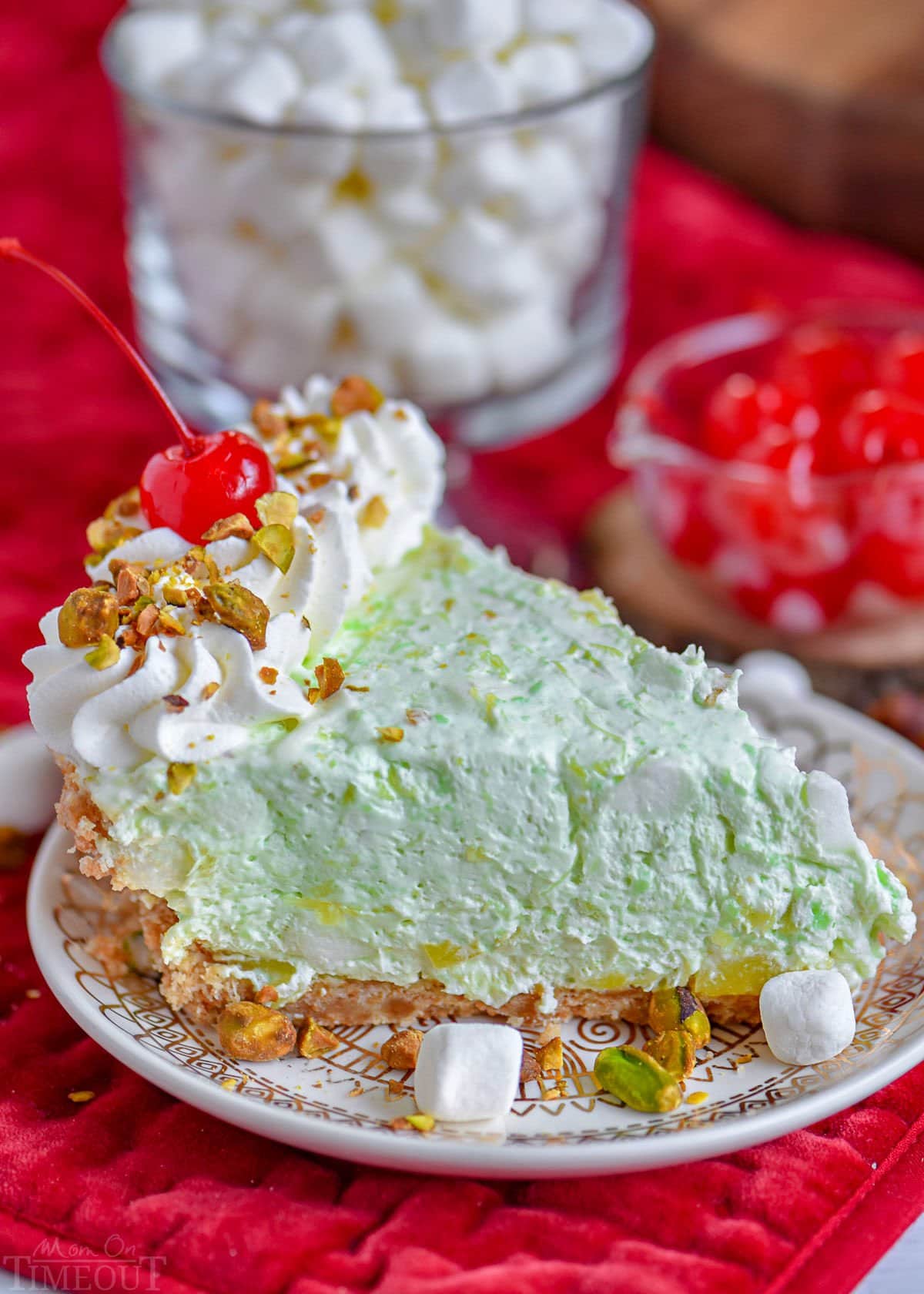 piece of pistachio pie on small white dessert plated garnished with freshly whipped cream, chopped pistachios and a maraschino cherry.