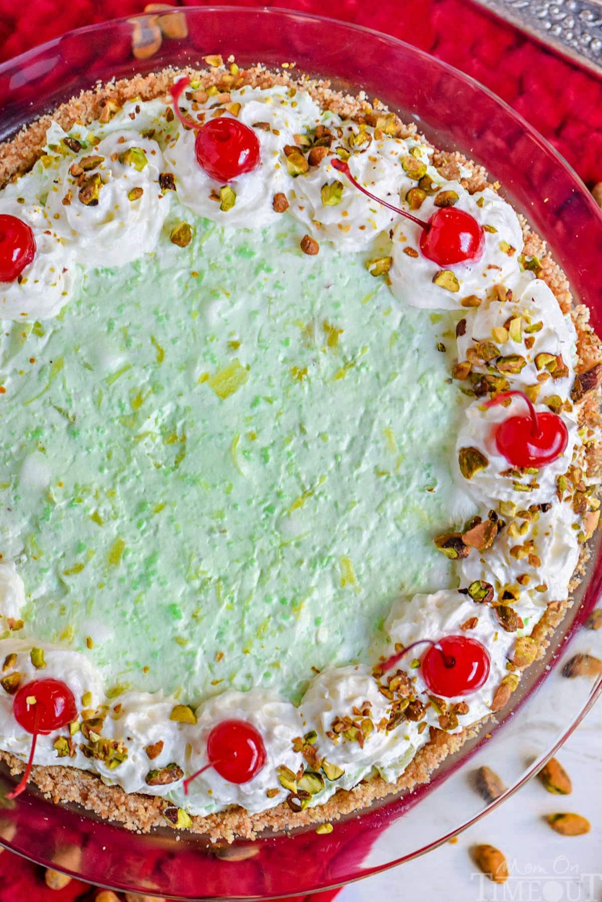 top down look at green pistachio pie decorated with whipped cream, pistachios, and maraschino cherries. pie is sitting on a bright red placemat.