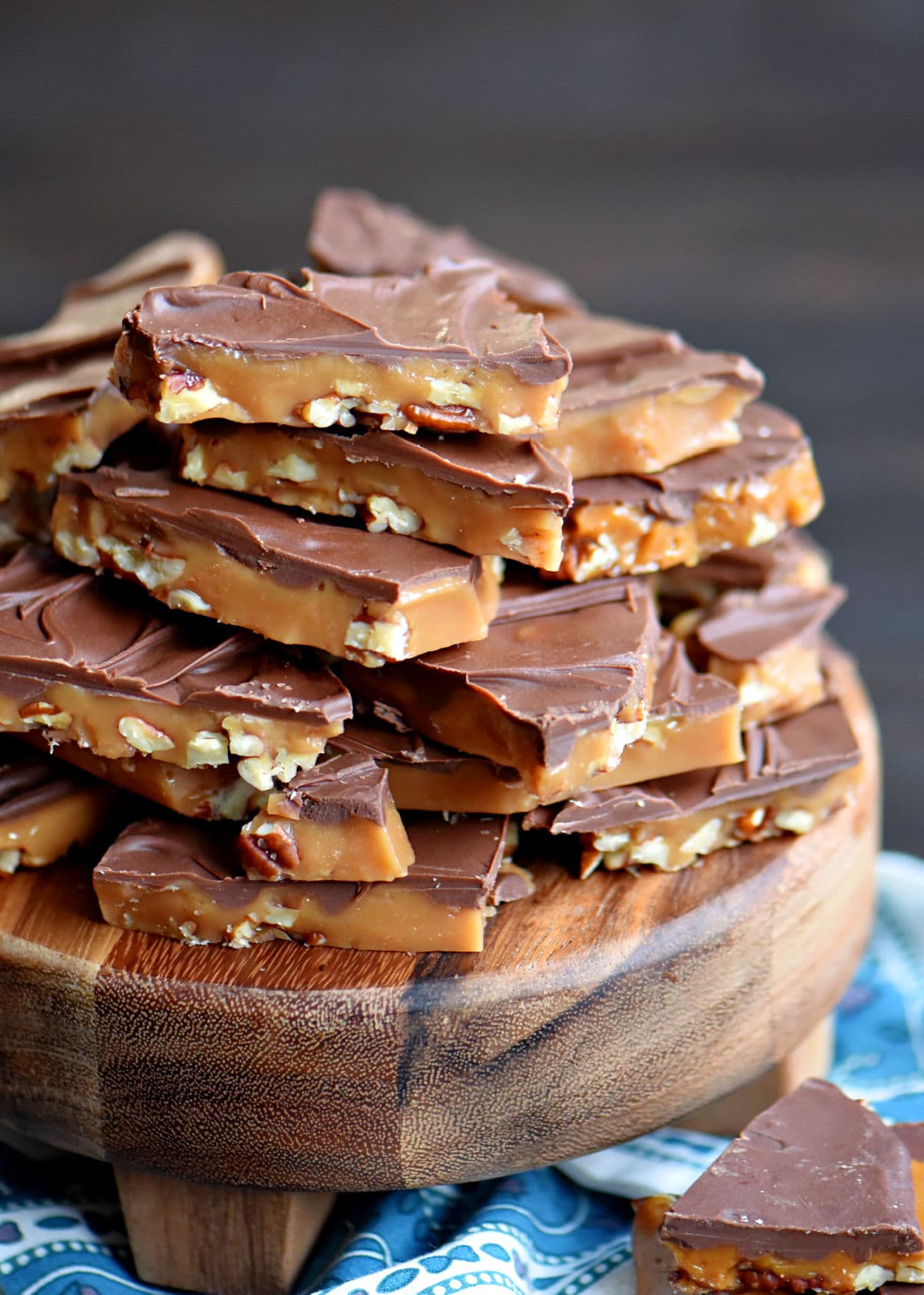 a large stack of toffee made with pecans and milk chocolate pile high on a dark wood round board.