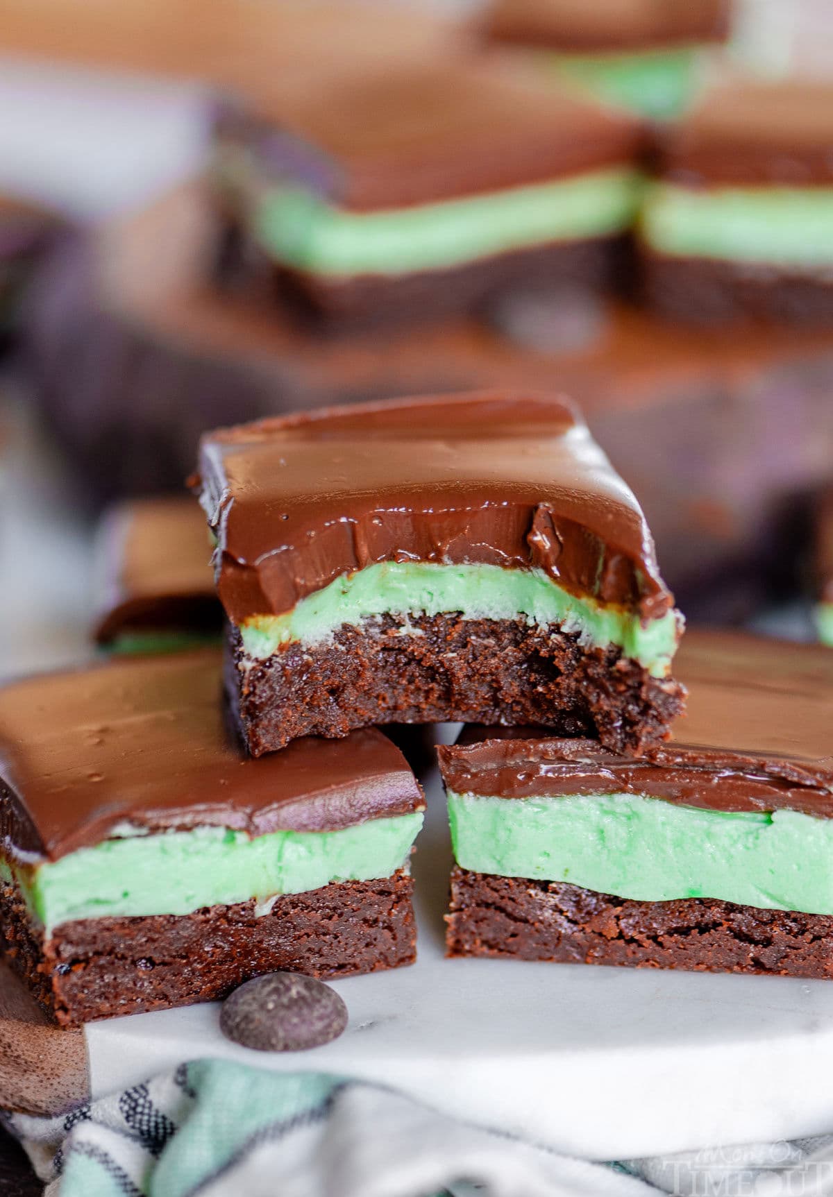 three mint brownies stacked in a pyramid. top brownie has bite taken out of it. more brownies can be seen in the background.