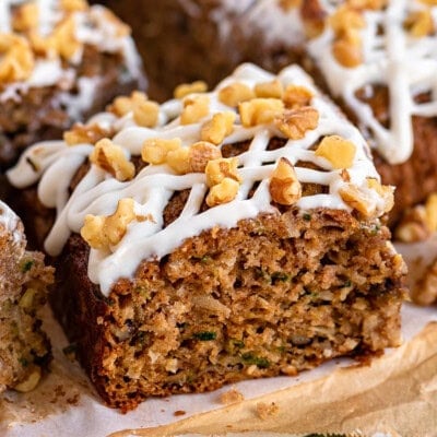 piece of zucchini spice cake on white board with the rest of the cut up snack cake behind it. The cake is topped with a cream cheese icing and walnuts.