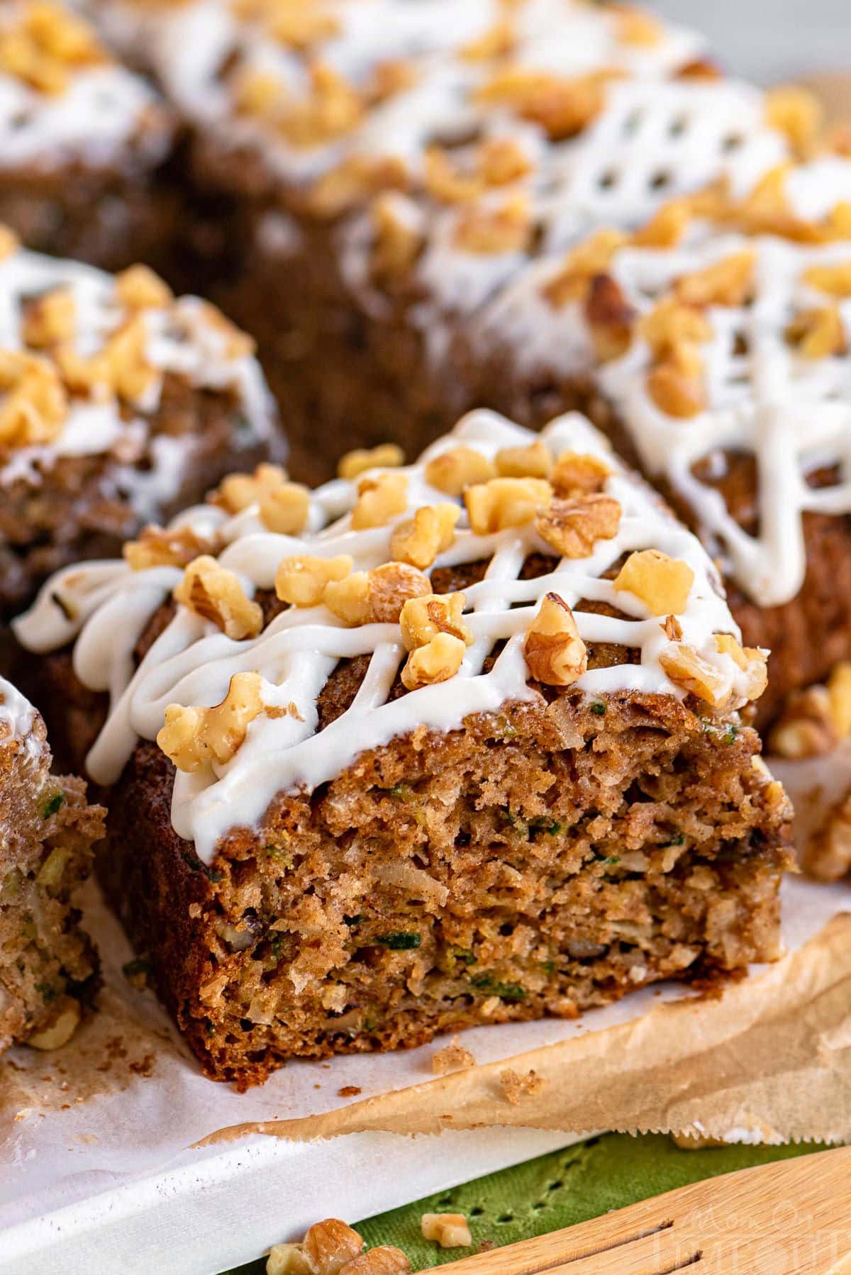 piece of zucchini snack cake on white board with the rest of the cut up snack cake behind it. The cake is topped with a cream cheese icing and walnuts.