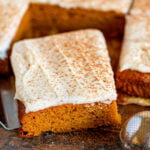 pumpkin bars cut into squares sitting on dark wood board with pumpkin pie spice dusted over the cream cheese frosting.