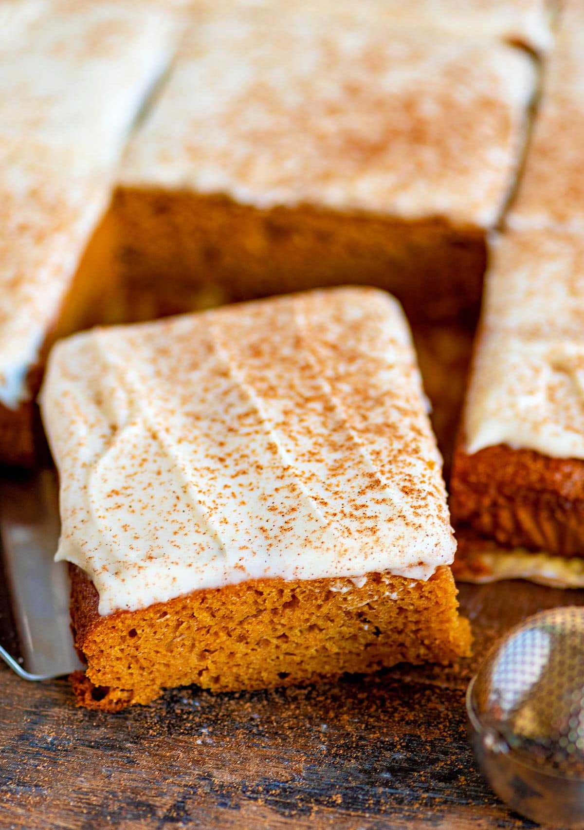 pumpkin bars cut into squares sitting on dark wood board with pumpkin pie spice dusted over the cream cheese frosting.