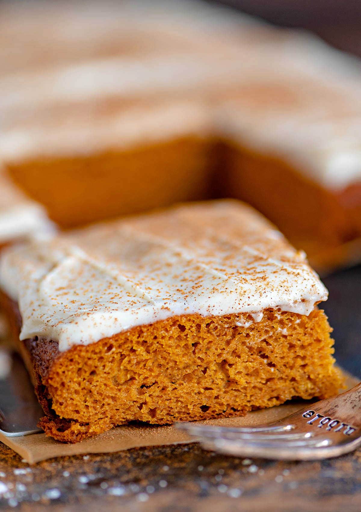 piece of pumpkin bars sitting on brown parchment on a dark wood surface with an antique fork sitting in front of it. The rest of the bars can be seen in the background.