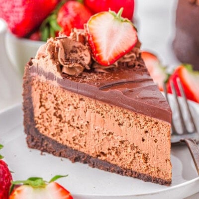 Slice of decadent chocolate cheesecake with an oreo crust and rich chocolate ganache on white round plate. Topped with chocolate shavings and fresh strawberries.