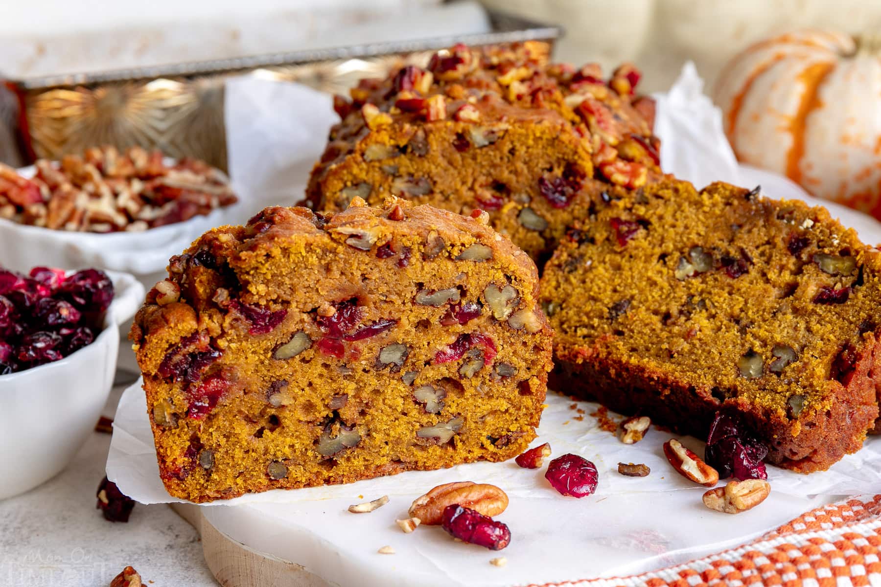 cranberry pecan pumpkin bread cut into slices and sitting on a piece of parchment with dried cranberries and pecans scattered about.