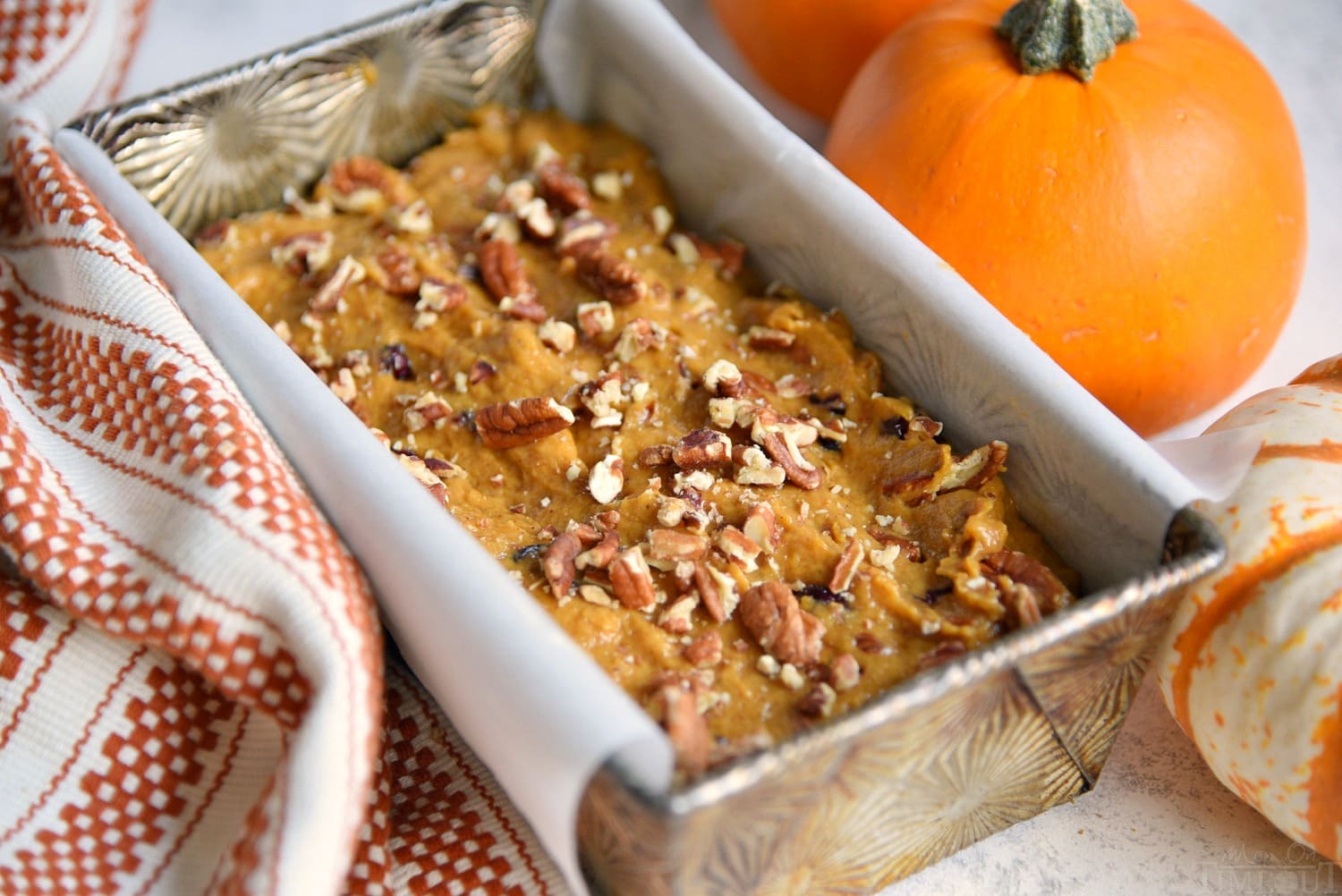 pumpkin batter in loaf pan that has been lined with parchment paper. fall colored towel sitting next to pan.