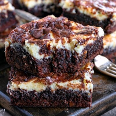 two cheesecake brownies stacked on each other on a dark wood board. more brownies can be seen in the background.
