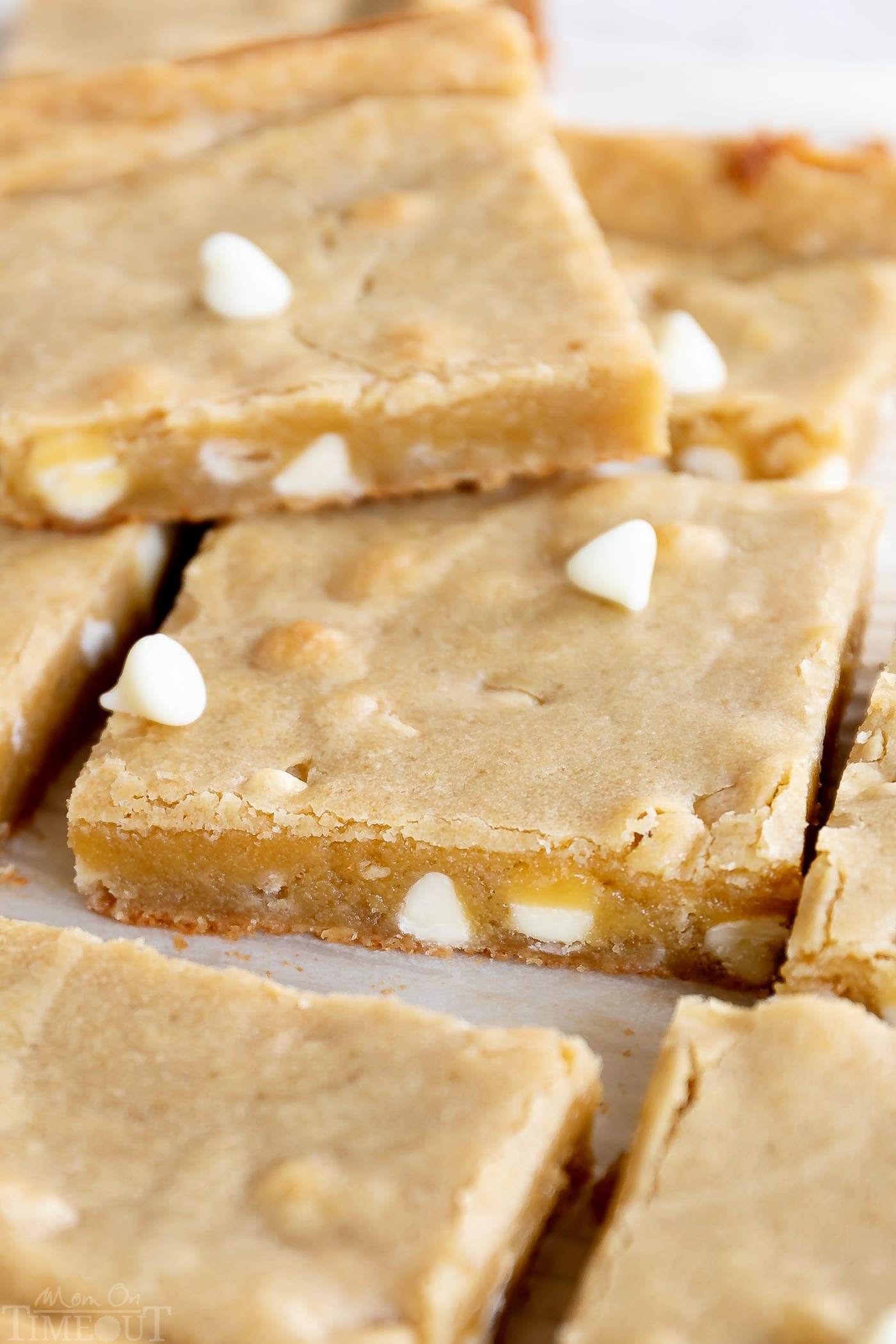 blondies made with white chocolate chips are cut into squares and sitting on white parchment. one blondie is sitting on top.