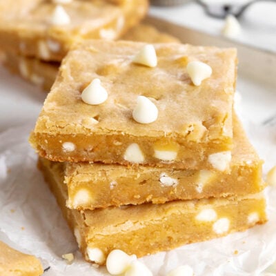 three blondie bars stacked with white chocolate chips sprinkled about sitting on white parchment with more bars in the background.