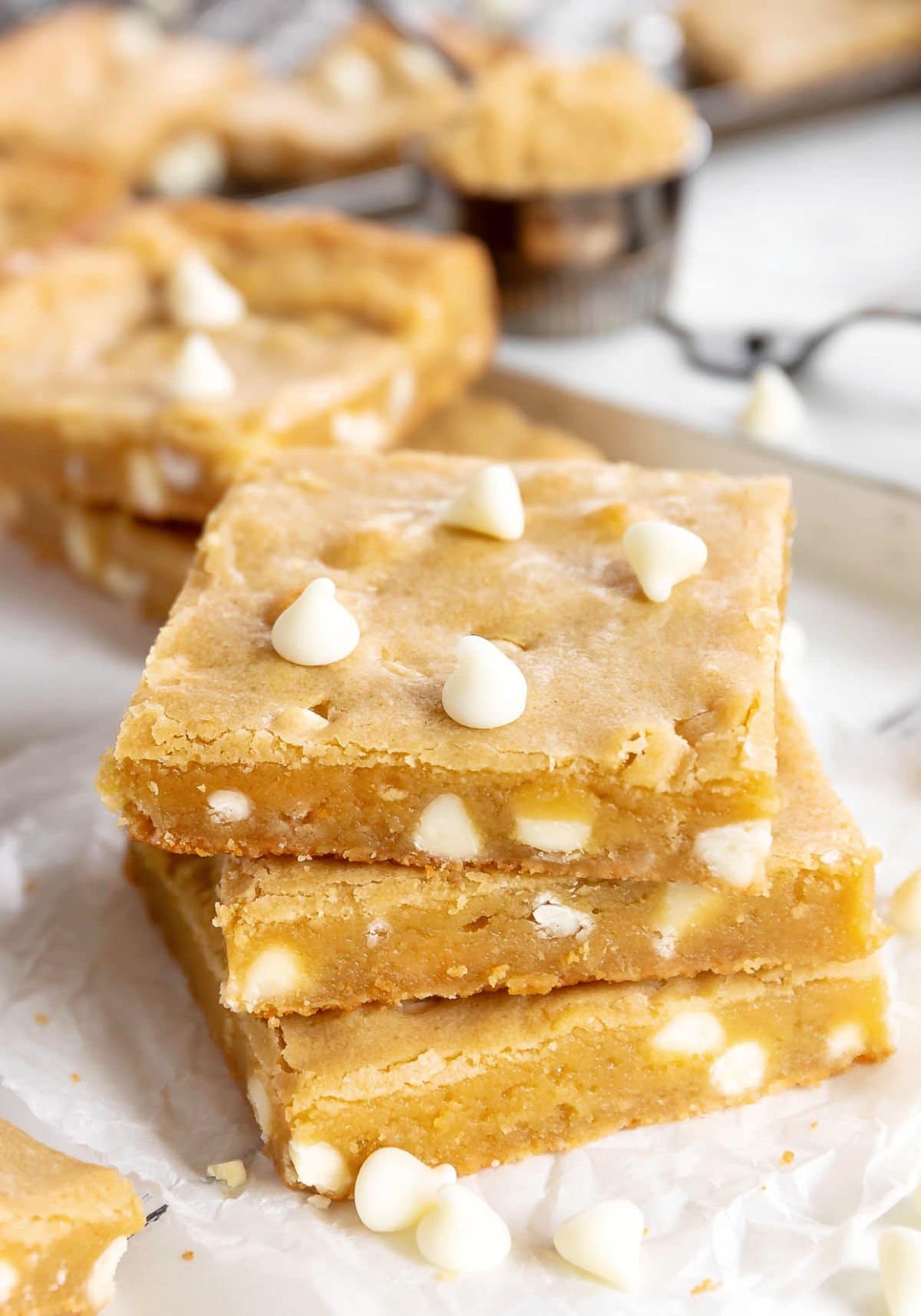 three blondie bars stacked with white chocolate chips sprinkled about sitting on white parchment with more bars in the background.
