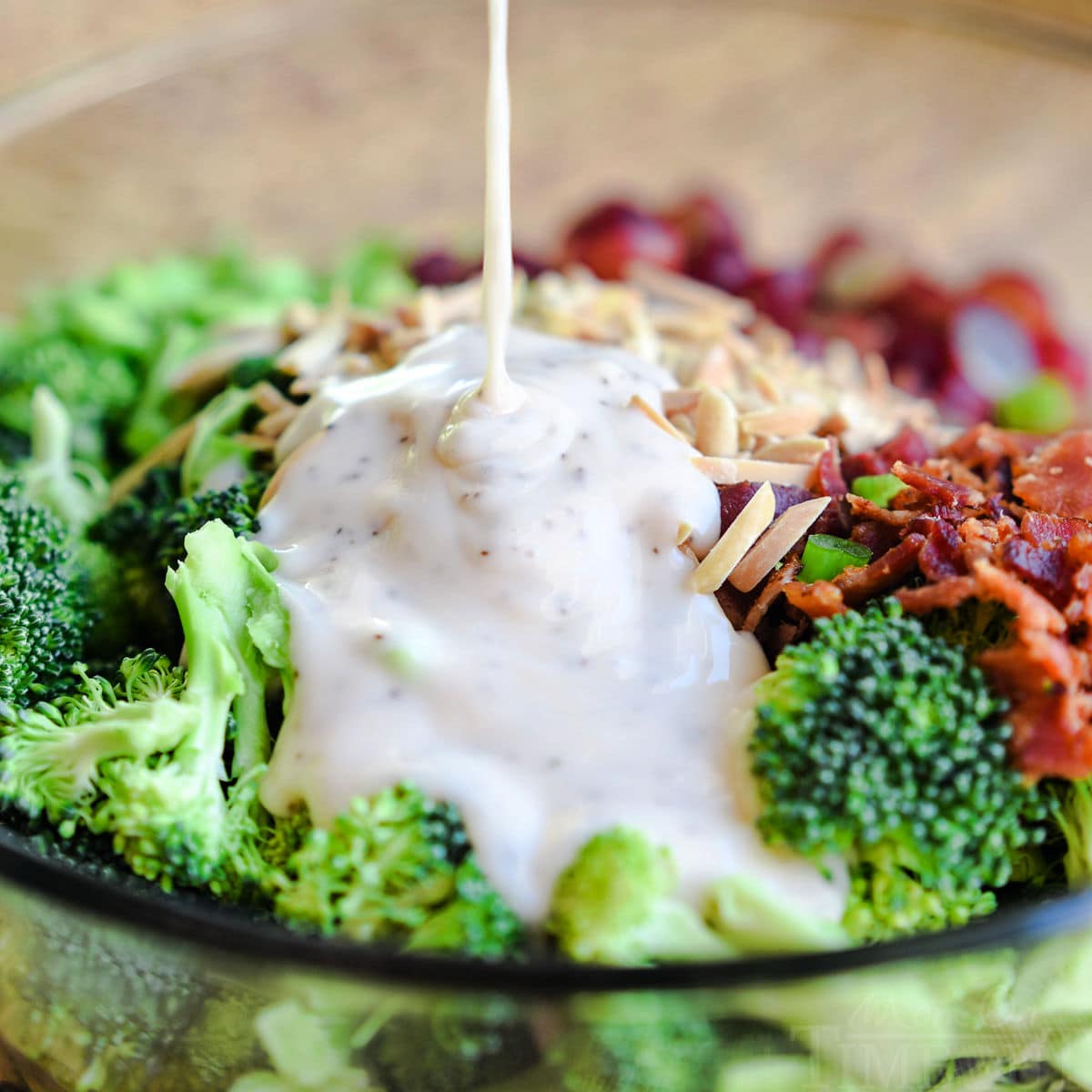 square image showing broccoli salad dressing being drizzled onto the rest of the ingredients in a large glass bowl.