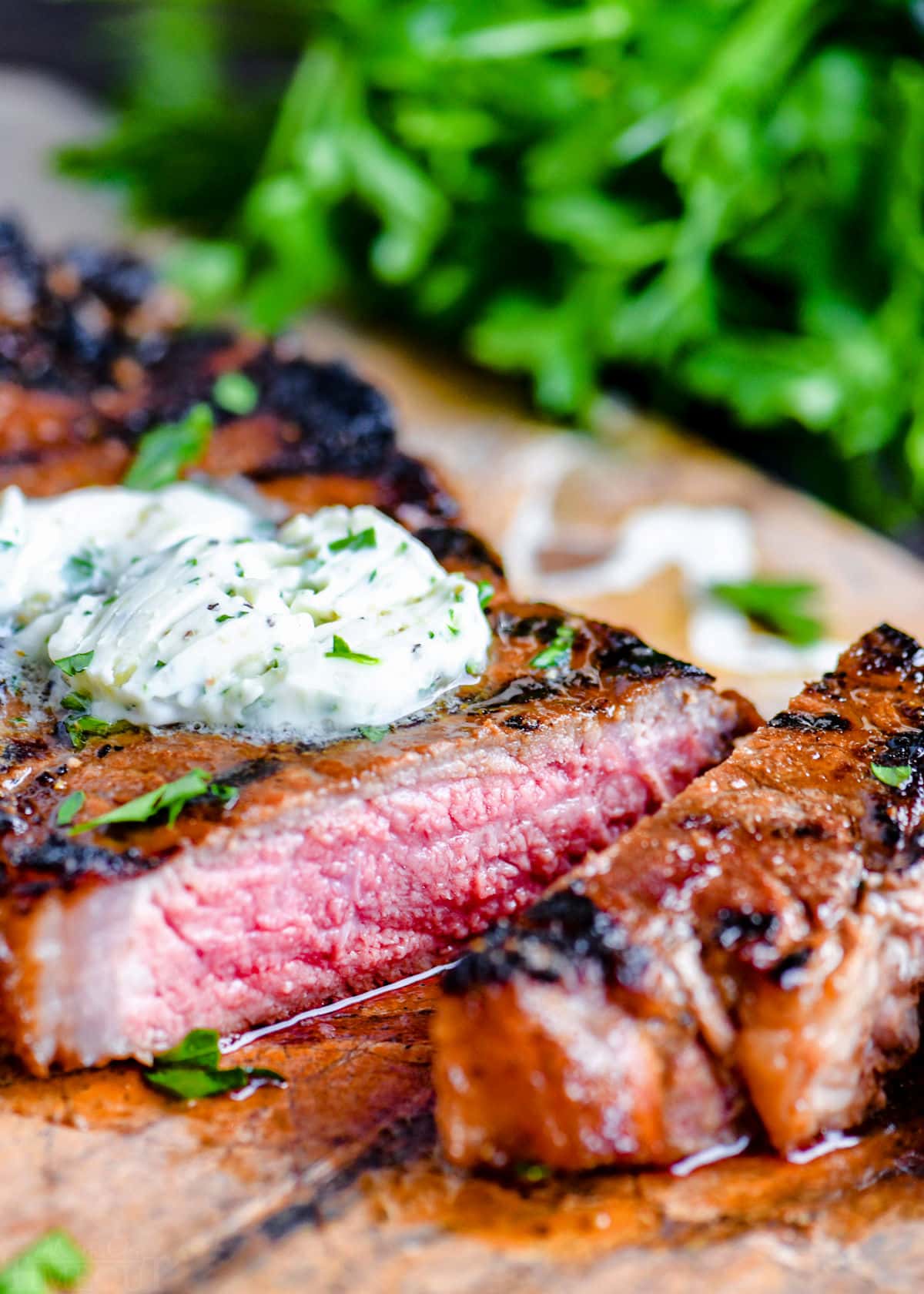 steak on cutting board with compound butter melting on top. one piece cut off of steak with fresh parsley in the background.