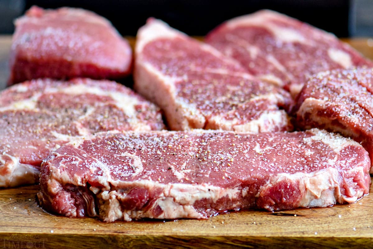 a variety of raw steaks seasoned with salt and pepper sitting on a wood board.