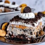 square piece of ice cream cake made with oreos sitting on silver plate and topped with whipped cream. silver baking dish in background with the rest of the cake in it.