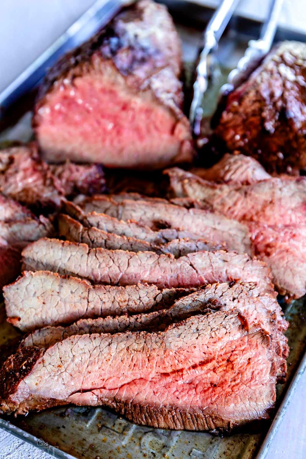sliced tri-tip with steak seasoning on a sheet pan with tongs