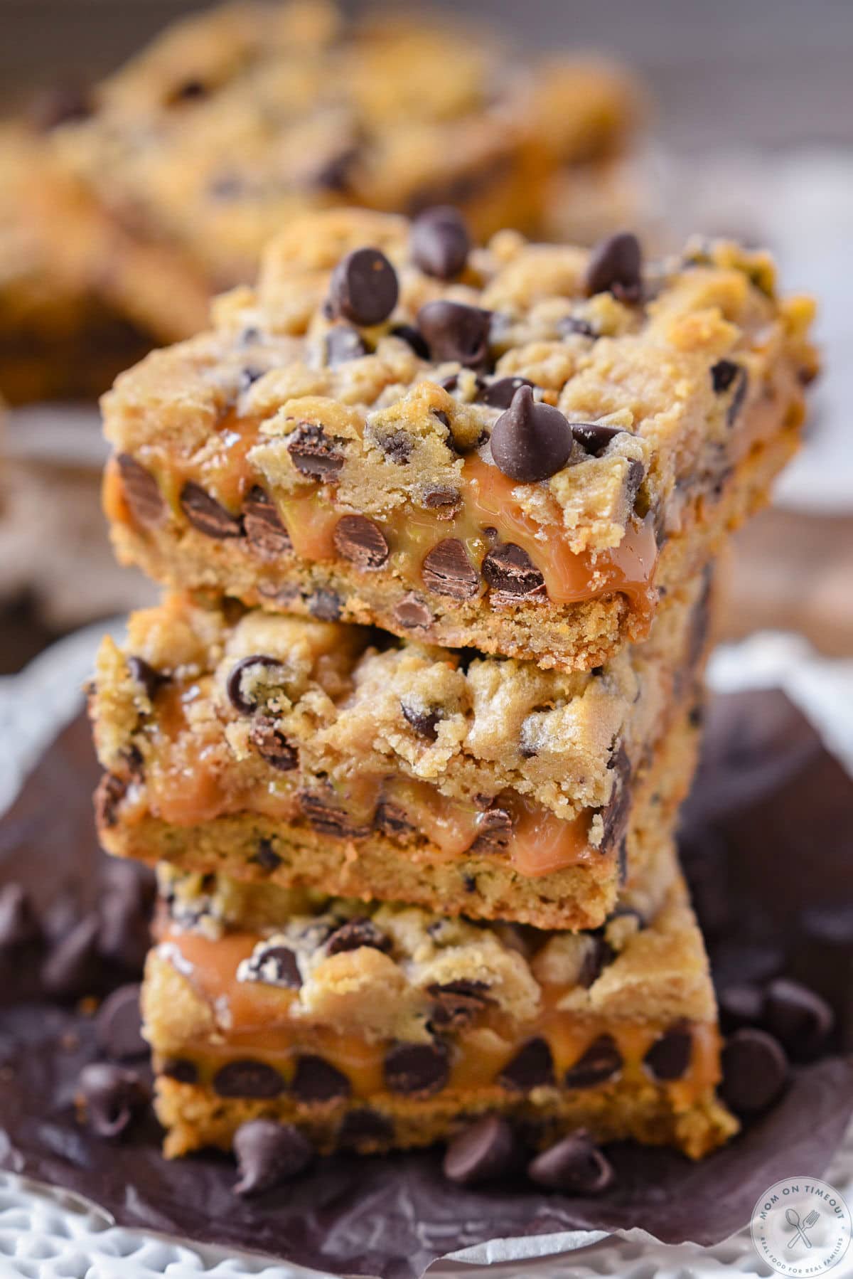 Stack of three carmelita bars with gooey caramel centers on small round dessert plate surrounded by chocolate chips. More bars can be seen in the background.