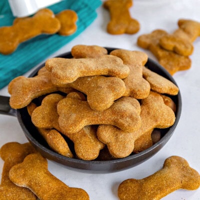 Homemade dog treats recipe cut into bone shapes and placed in large black bowl. More treats are scattered around the bowl.
