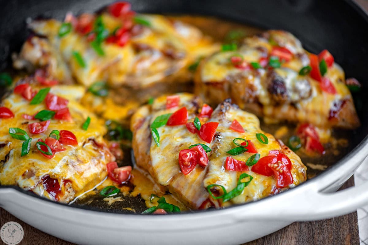 Wide shot of four chicken breasts in skillet topped with bbq sauce, bacon, cheese and fresh green onions and tomatoes. 