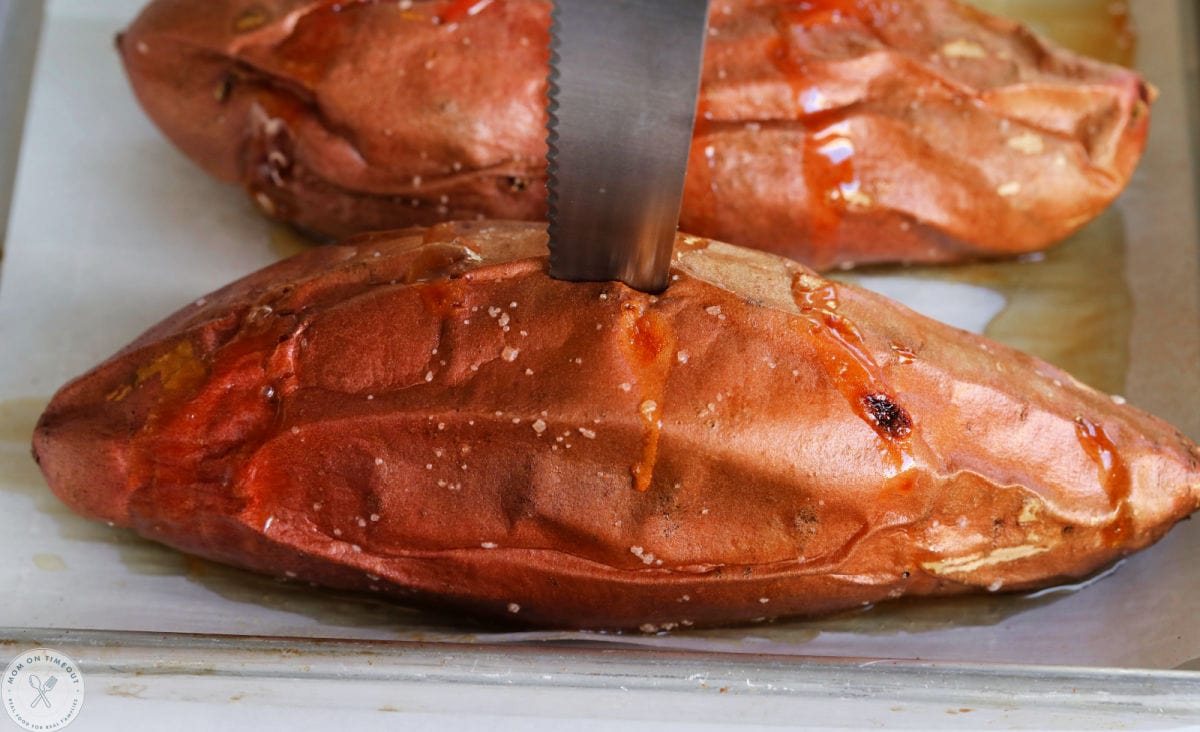 Baked sweet potatoes in oven fully cooked with knife inserted to show doneness. Sweet potato is on a parchment lined baking sheet.
