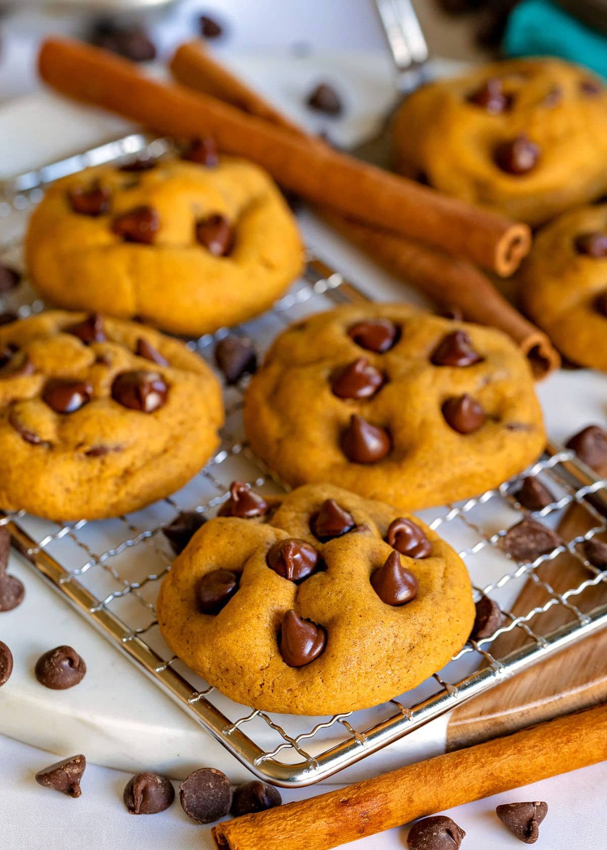 four pumpkin chocolate chip cookies sitting on wire rack with cinnamon sticks and chocolate chips scattered near them.