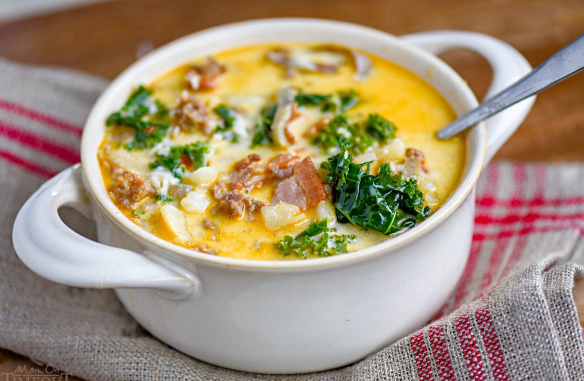 white bowl with handles filled with zuppa toscana soup and topped with cheese. Kale, sausage and bacon can be seen in the soup.