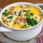 white bowl with handles filled with zuppa toscana soup and topped with cheese. Kale, sausage and bacon can be seen in the soup.