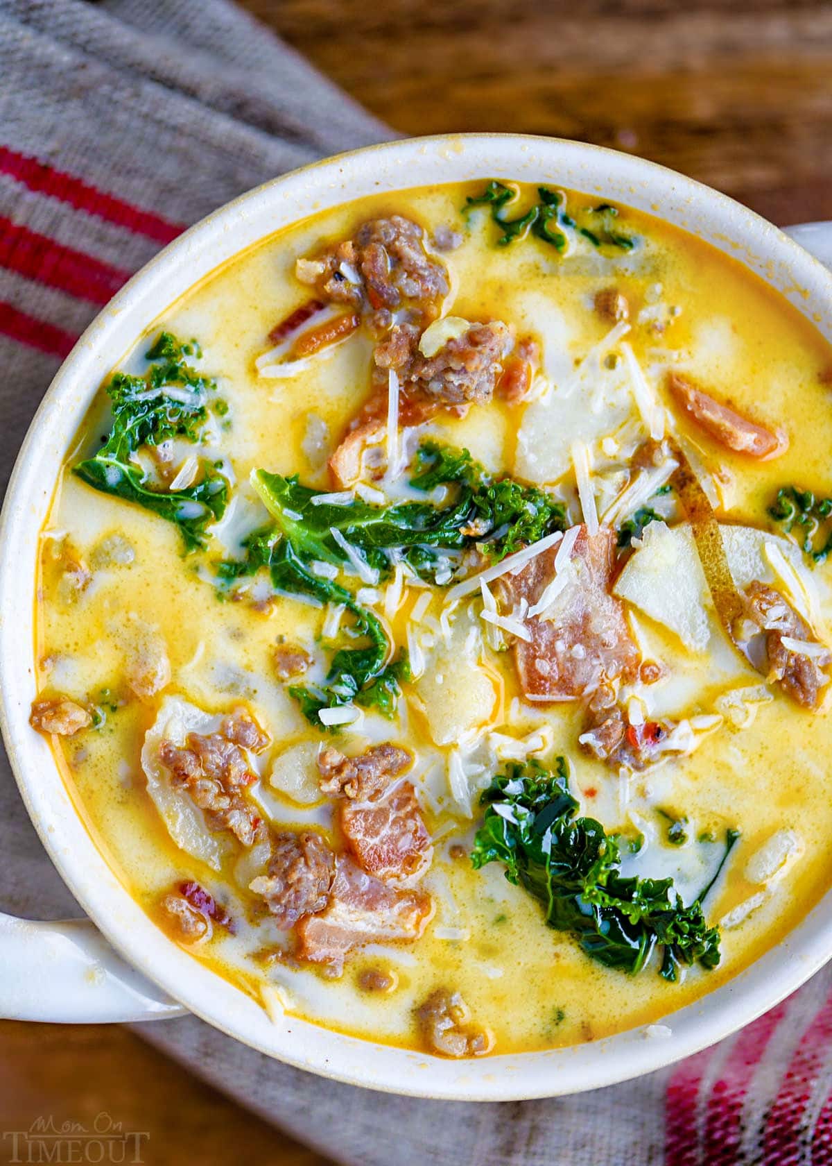 top down view of a bowl of zuppa toscana soup in a white bowl with handles. Soup is topped with Parmesan cheese and is sitting on a brown and red linen napkin.