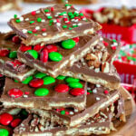 cut up pieces of christmas crack recipe on wood stand piled high with nuts sprinkles and mms. more pieces of the toffee seen in the background.