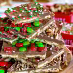 cut up pieces of christmas crack recipe on wood stand piled high with nuts sprinkles and mms. more pieces of the toffee seen in the background.
