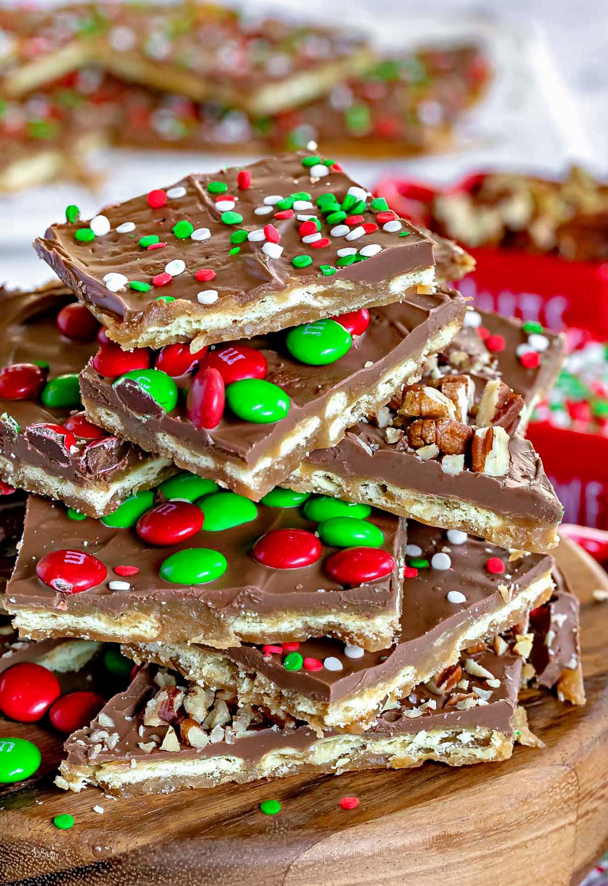 cut up pieces of christmas crack recipe on wood stand piled high with nuts sprinkles and mms. more pieces of the toffee seen in the background. 