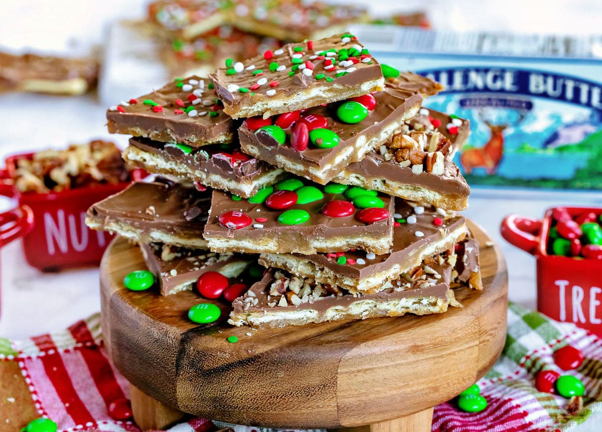 over a dozen large squares of saltine toffee sitting on round wood serving board sitting on a Christmas towel. Little red ceramic containers are in the background filled with candies and nuts.